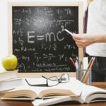 Woman writing physics equations on a blackboard with books and an apple on the desk.
